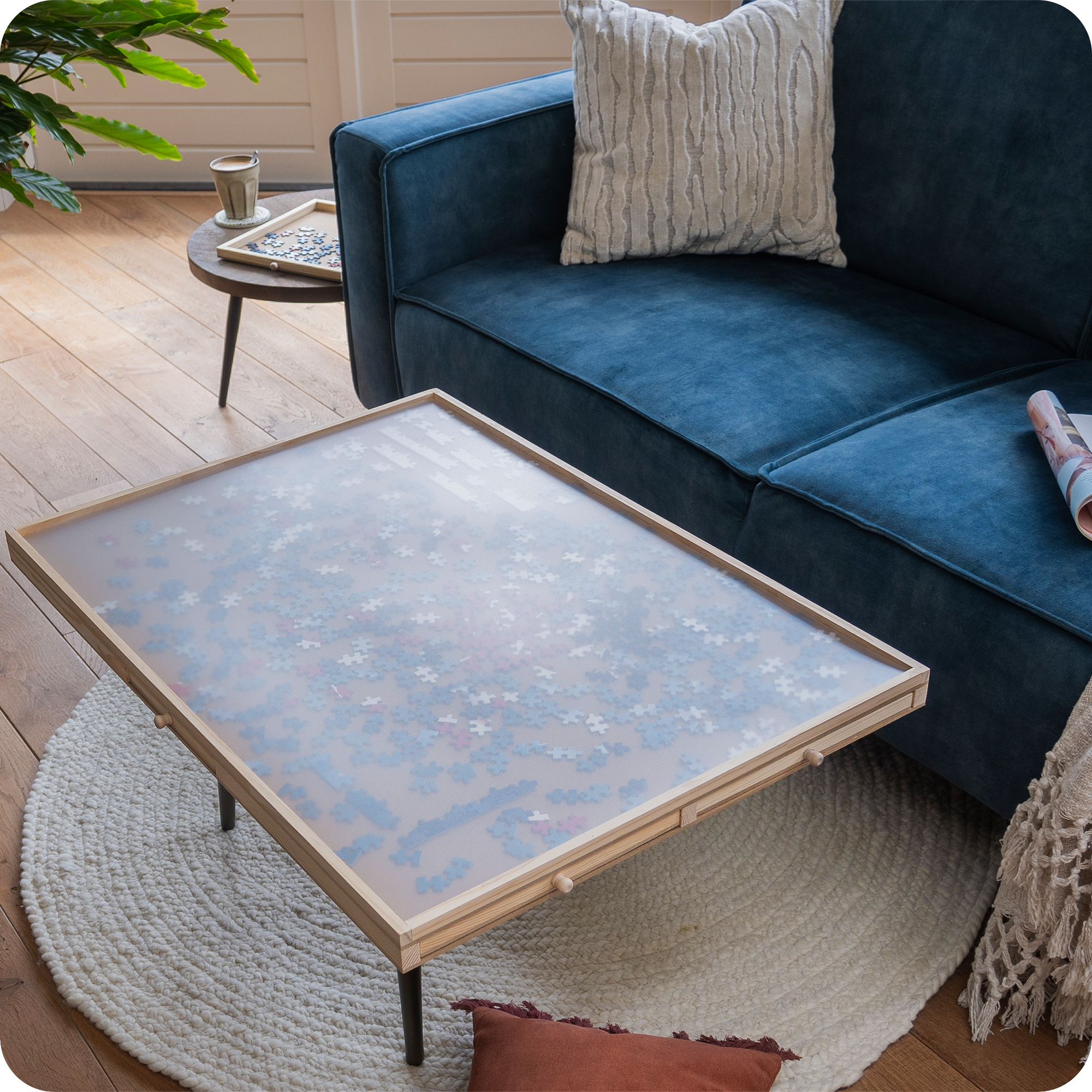 Een landelijke woonkamer met een blauwe bank en landelijke kussens en plaid. Op de salontafel staat de puzzelplank met een doorzichtige afdekplaat waardoor de puzzelstukjes worden beschermd.